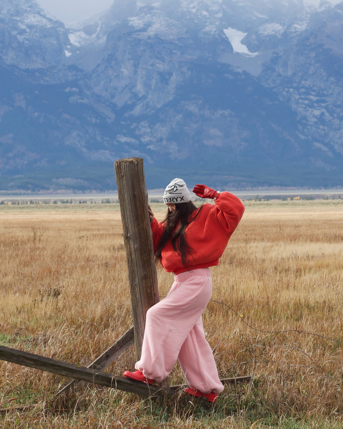 A person in a red sweatshirt, pink pants, and a white beanie stands in a grassy field with large mountains in the background, leaning against a wooden fence post.