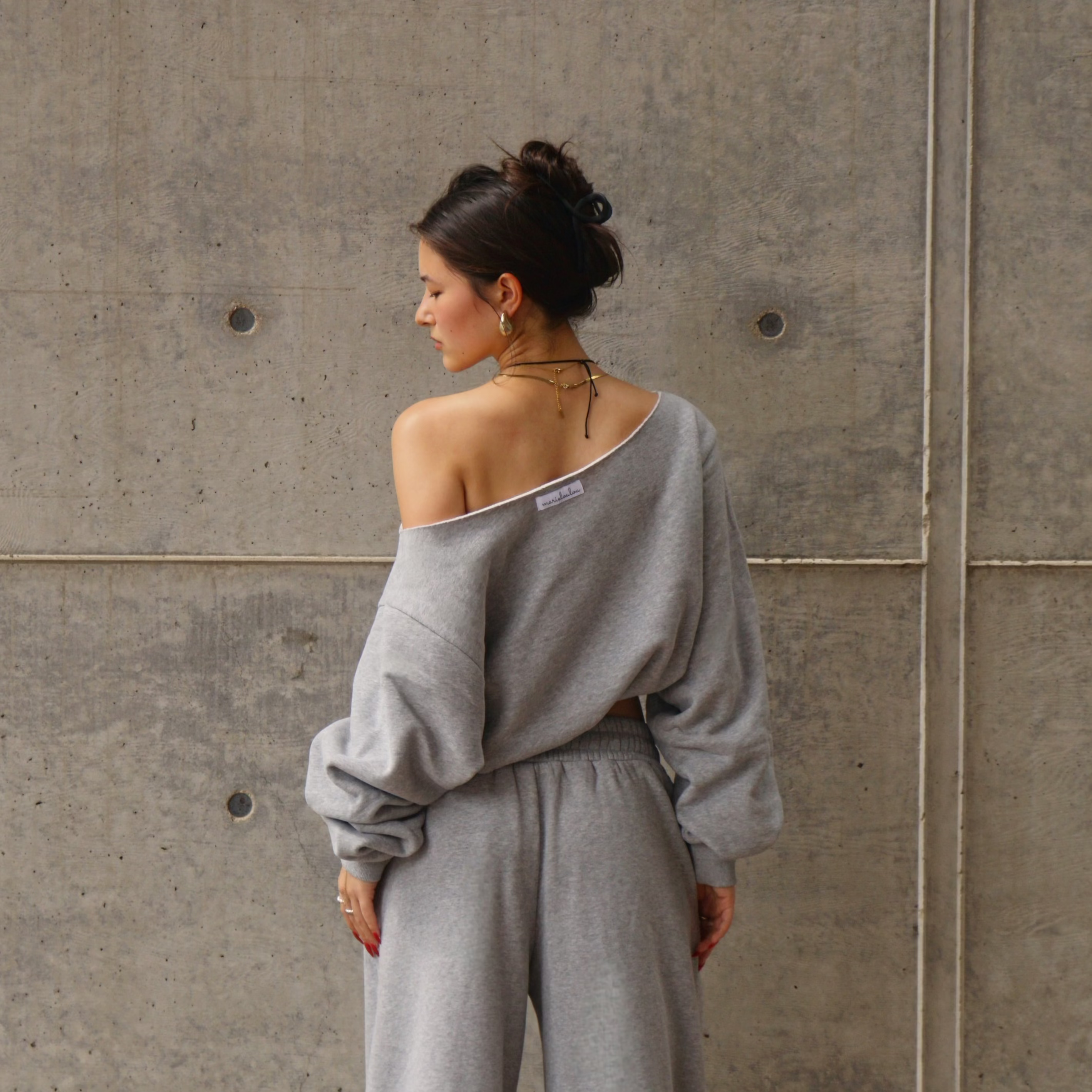 Woman wearing a gray off-shoulder sweatshirt and sweatpants against a concrete wall.