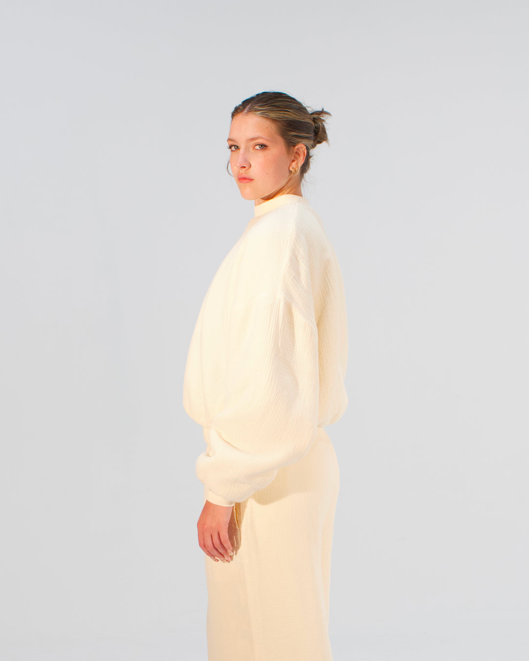 A model wearing a cream-colored, textured knit two-piece outfit consisting of a mock-neck long-sleeve top and matching trousers, shown from a side-back profile against a light gray studio background.