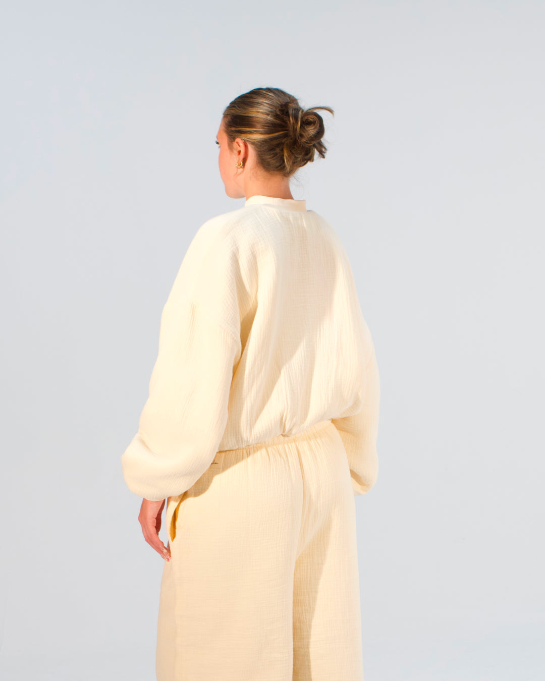 A woman shown from the back wearing a cream-colored linen jumpsuit against a light gray studio background.