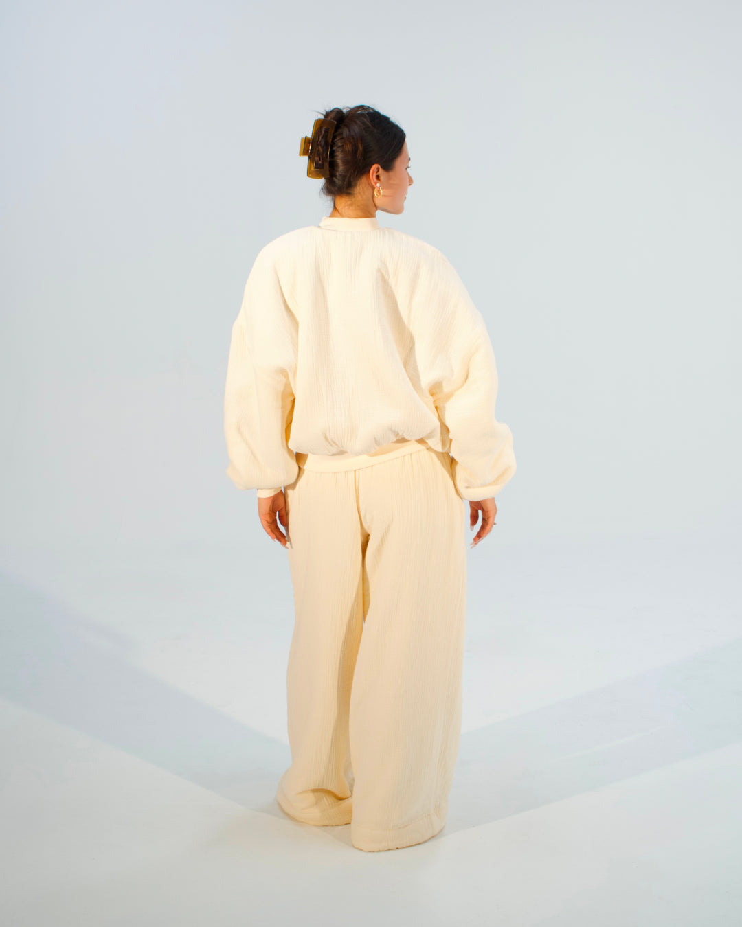 A model viewed from behind wearing a cream-colored two-piece lounge set consisting of a long-sleeve sweatshirt and matching wide-leg trousers, set against a light gray studio background.
