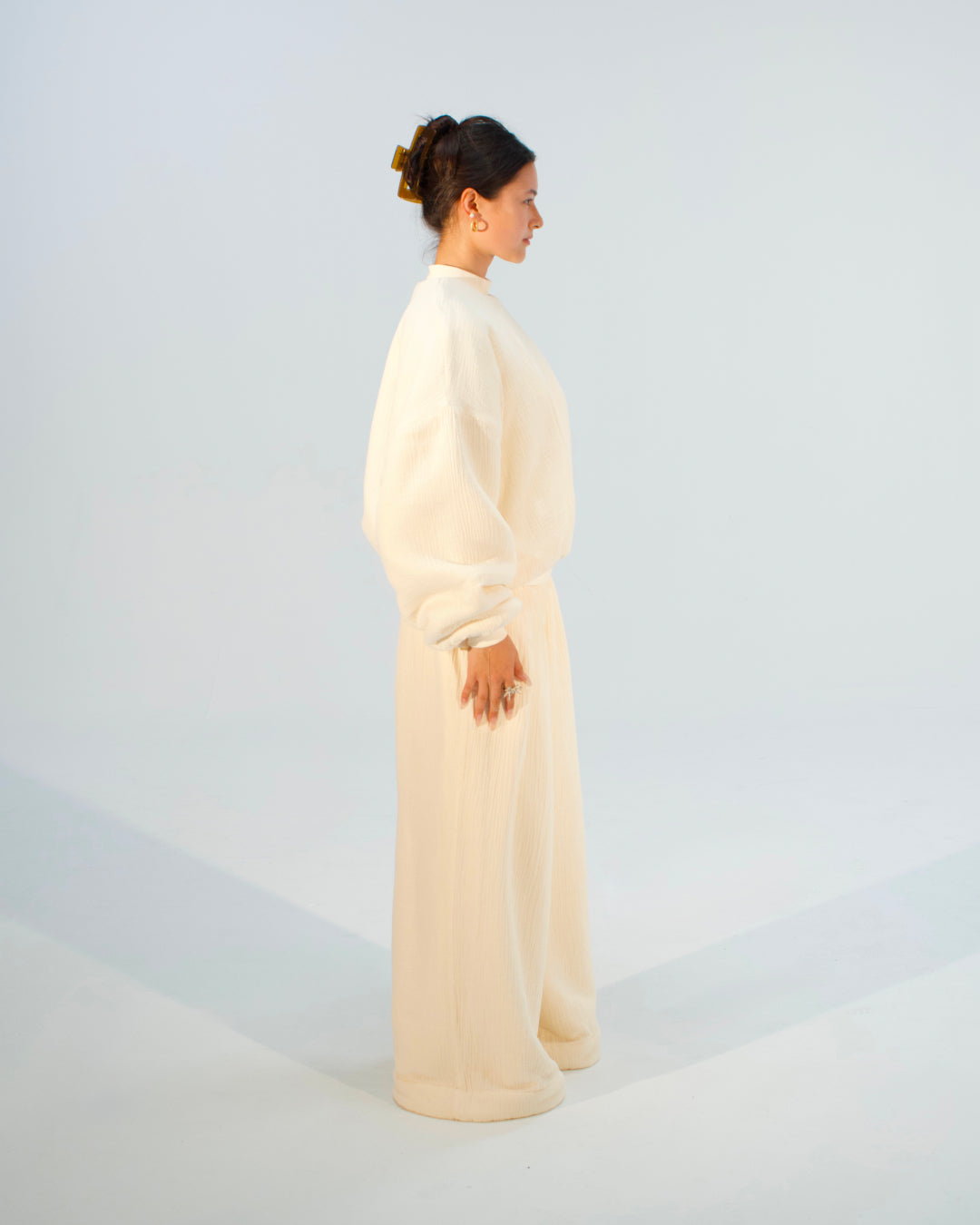 A woman stands in profile wearing a loose, high-neck, long-sleeve cream or off-white outfit with wide legs, photographed against a clean light gray studio background.