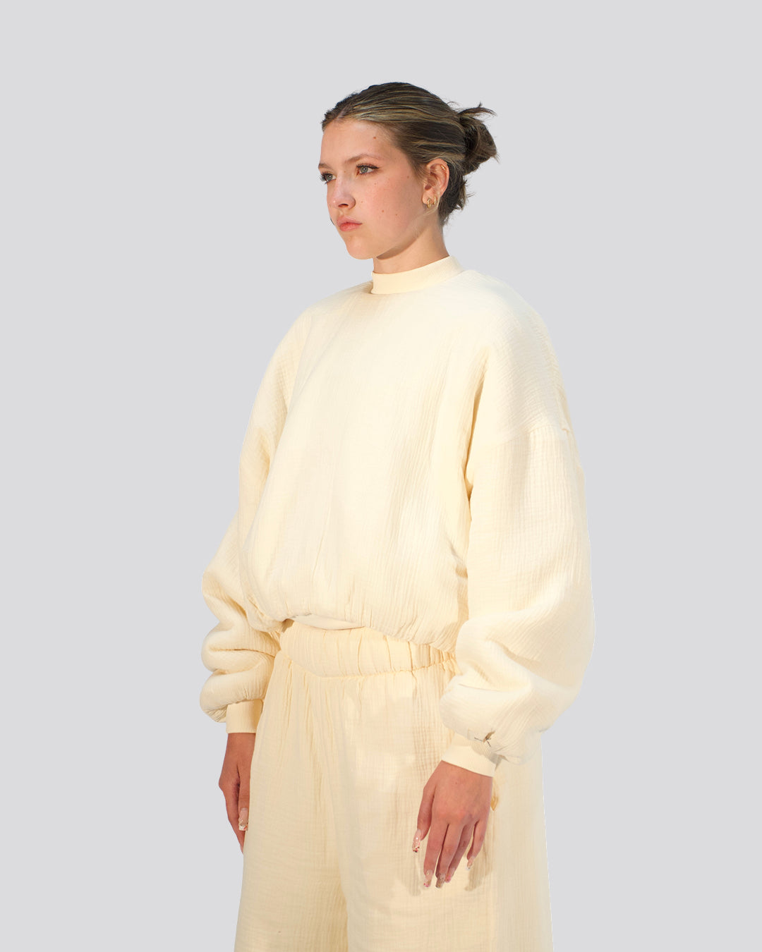 A model wearing a cream-colored, textured long-sleeve mock-neck top and matching wide-leg trousers, photographed against a solid light gray background.