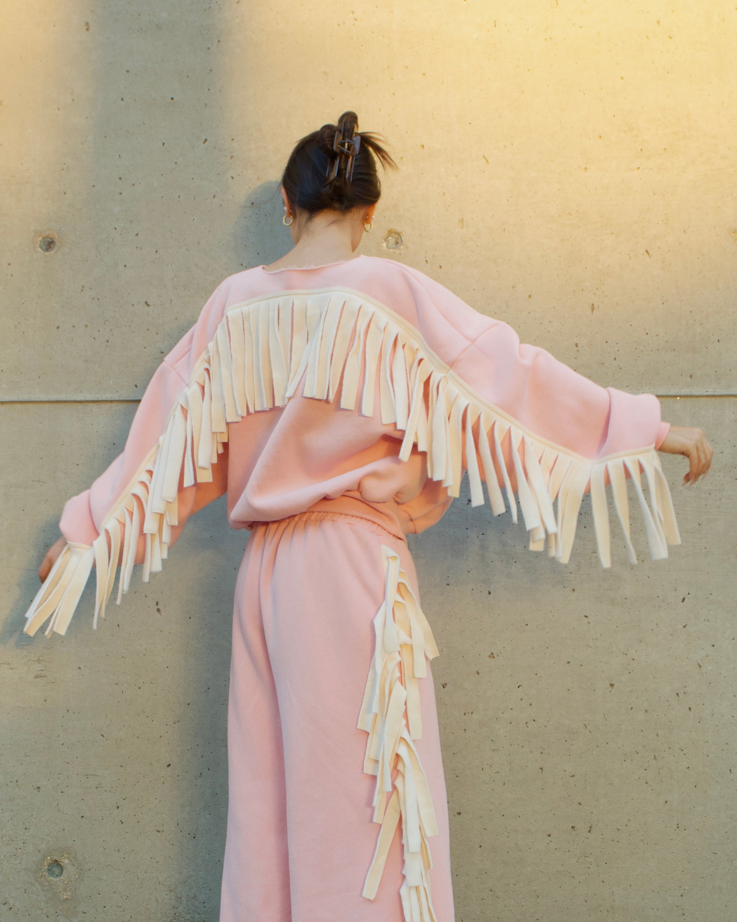 A model stands with their back to the camera wearing a light pink two-piece outfit with dramatic cream fringe details on the shoulders and sides, set against a concrete wall with warm sunlight.