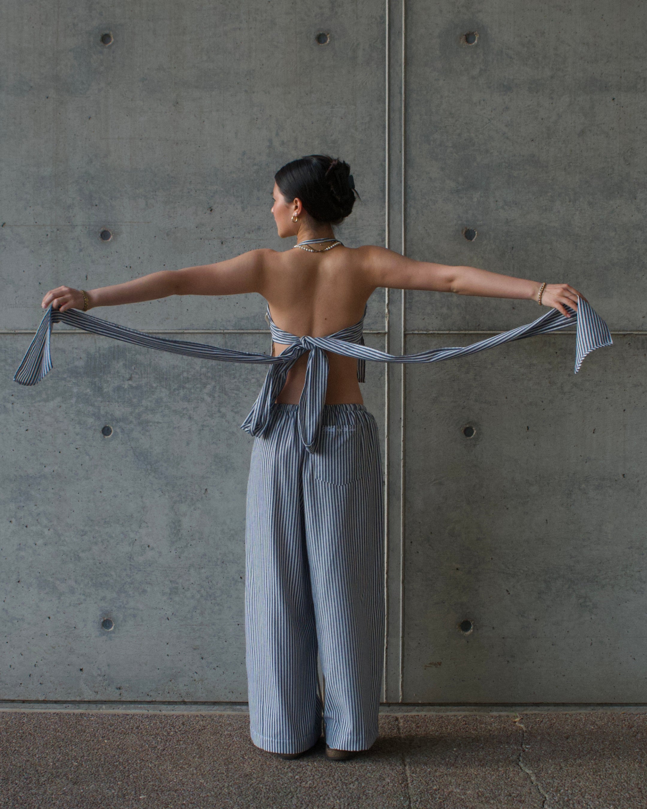 Woman in a blue striped napkin top and same color long wide leg pants standing against a concrete wall