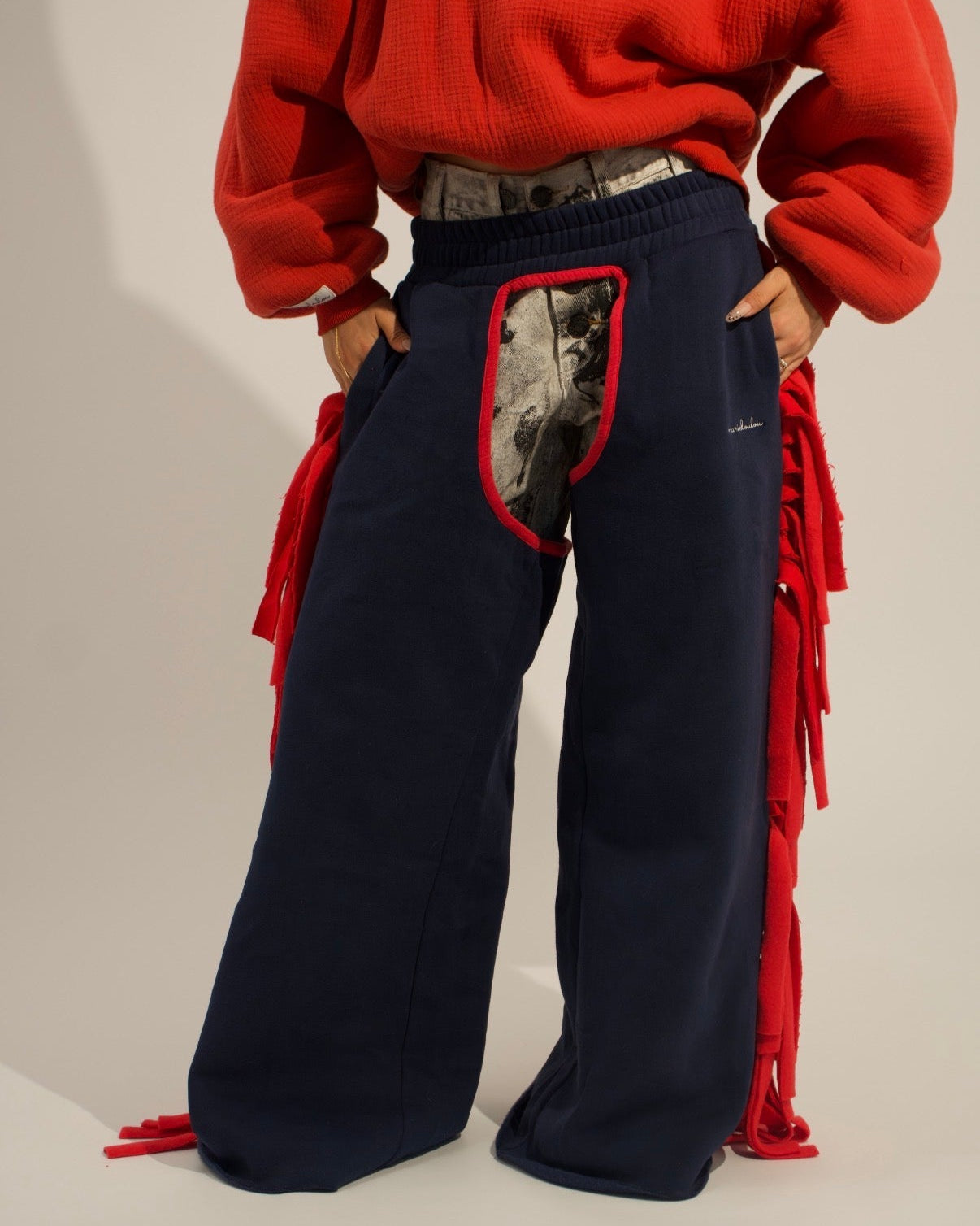 A person wearing oversized navy wide-leg sweatpants with red fringe details and a red-and-black patch pocket, paired with a bright red sweatshirt against a plain light beige studio background.