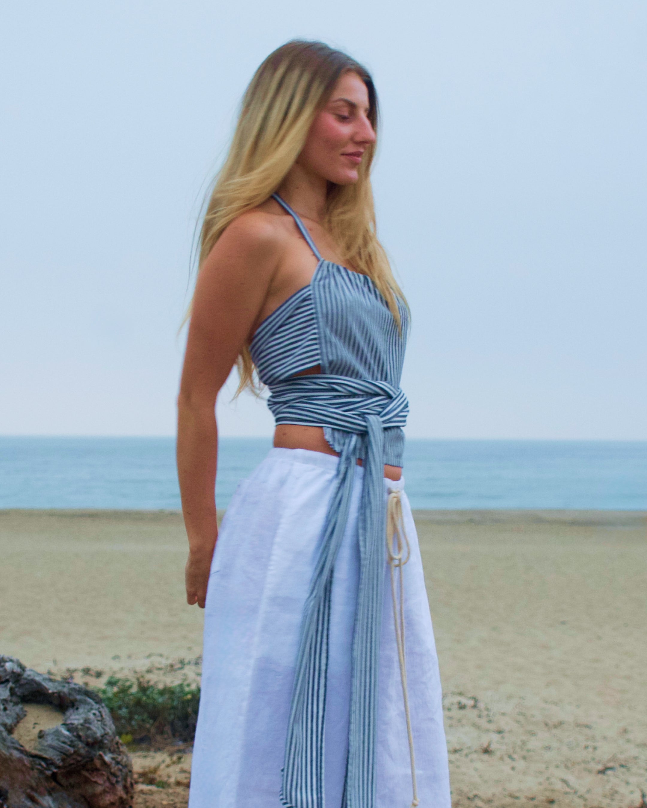 Woman in a Marieloulou Linen Stripe Napkin Top and  white Earth Linen pants standing on a beach with ocean in the background