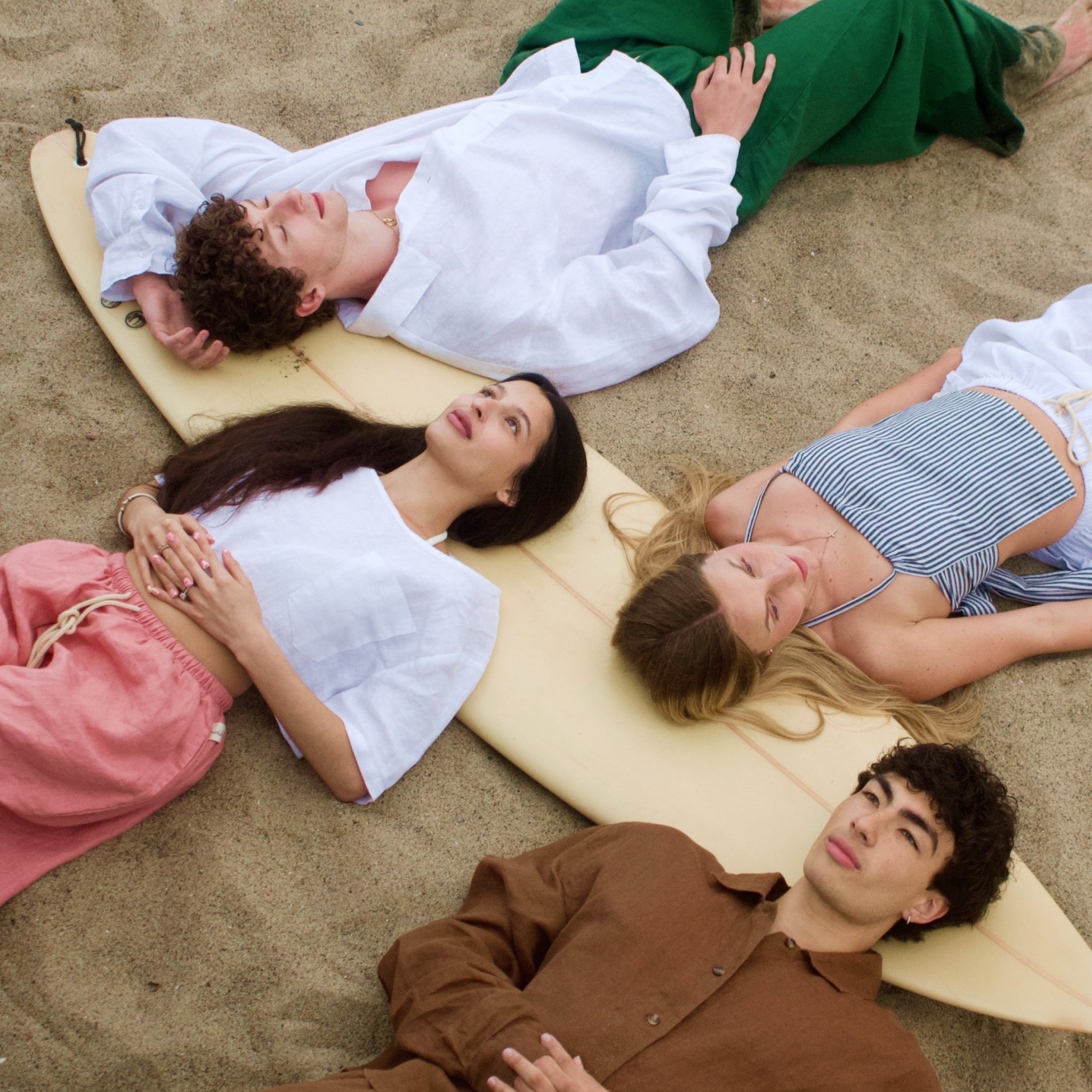 Four people lying on a surfboard on a sandy beach wearing Marieloulou Earth Linen Sets